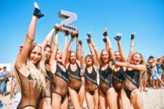 many girls in swimming suits on seashore holding festival logo