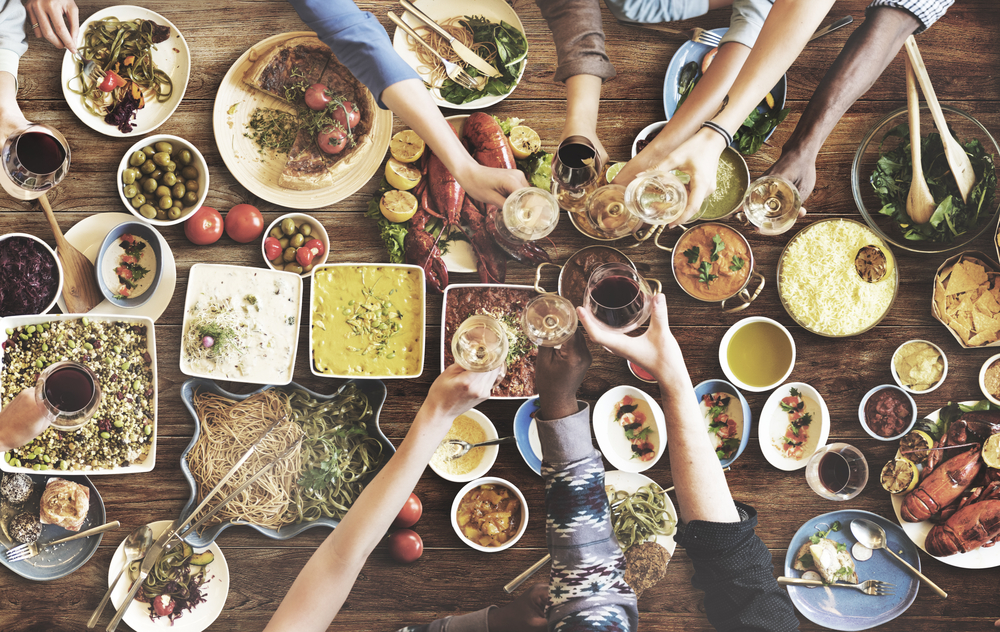 hands holding glasses over served table with many dishes